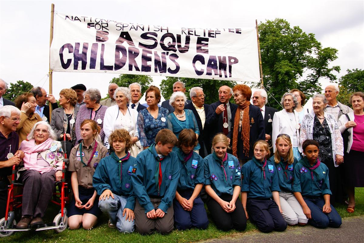 Varios ancianos posan con un grupo de niños y niñas scouts bajo un cartel que reza "BAsque children's camp"
